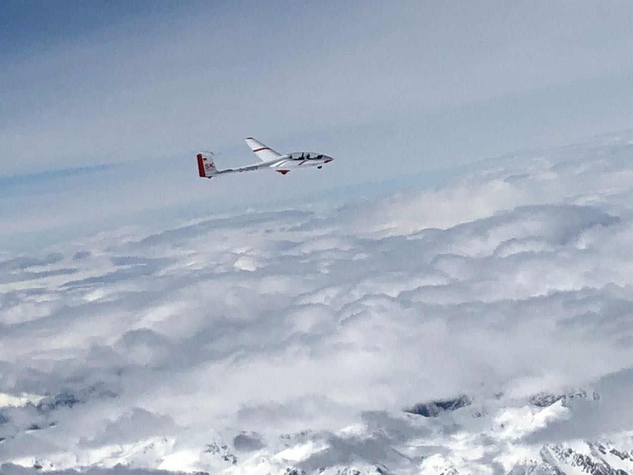 Eine ASK 21 mit roten Warnmarkierungen oberhalb der fast geschlossenen Wolkendecke. Im Vorderungd sieht man schneebedeckte Berggipfel durch die Wolkenlücken.